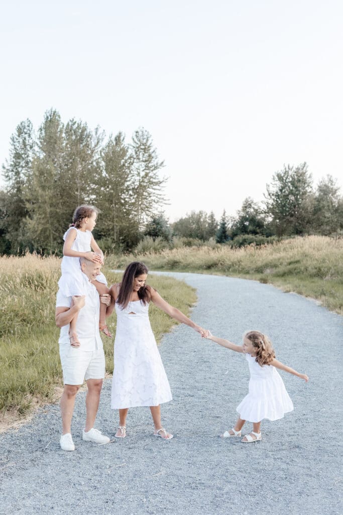 Family of four walking hand in hand down a path, parents in white with two daughters in matching dresses — relaxed family session