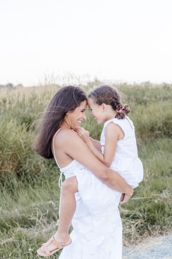 Mom holding young daughter in white dress, both smiling during a golden-hour family session in Abbotsford with Meliza Orellana Photography