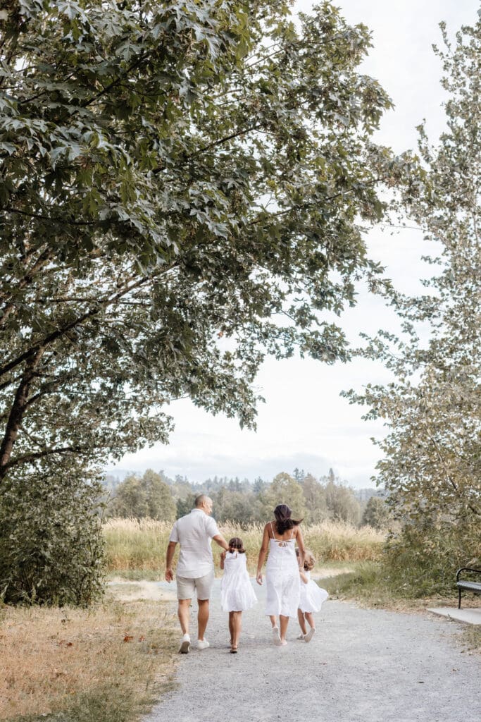 Family of four walking hand in hand down a path, parents in white with two daughters in matching dresses — relaxed family session