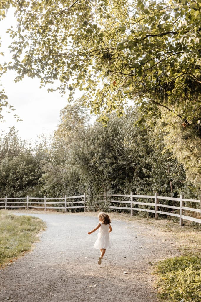 Young girl skipping down a gravel path surrounded by tall grass during sunset — outdoor family photo by Vancouver family photographer Meliza Orellana