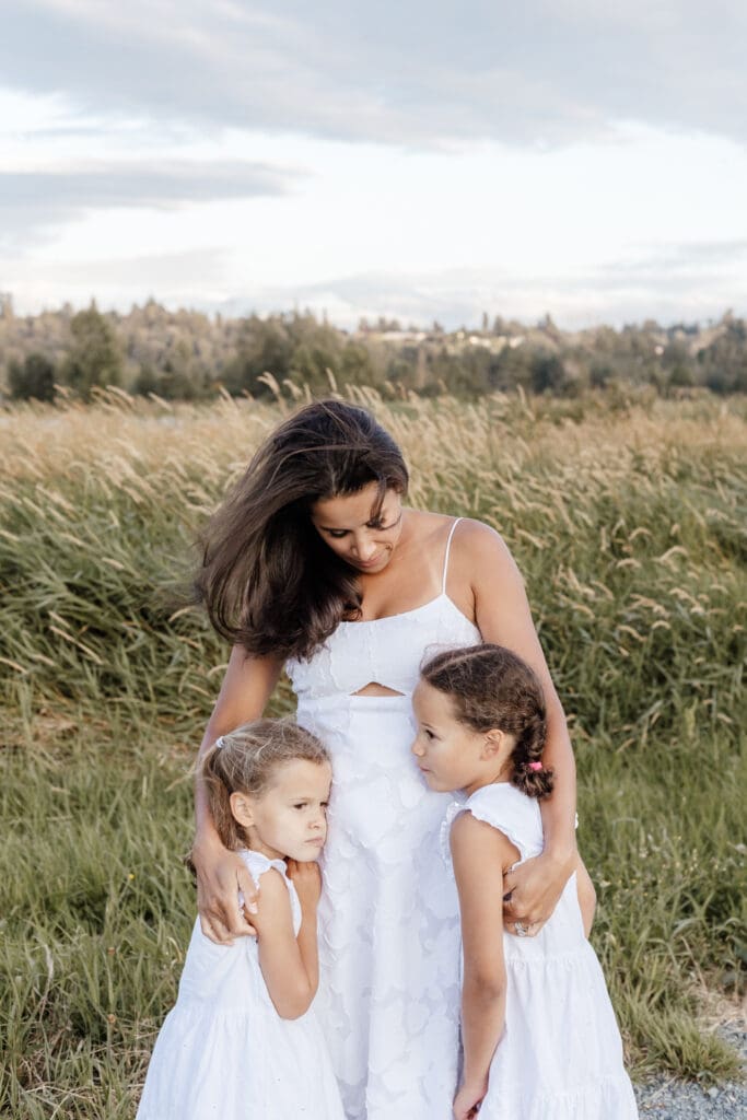 Mother standing in field of grass with two daughters, one on each side — timeless family portrait captured outdoors in Willband Creek Park in Abbotsford by Meliza Orellana Photography