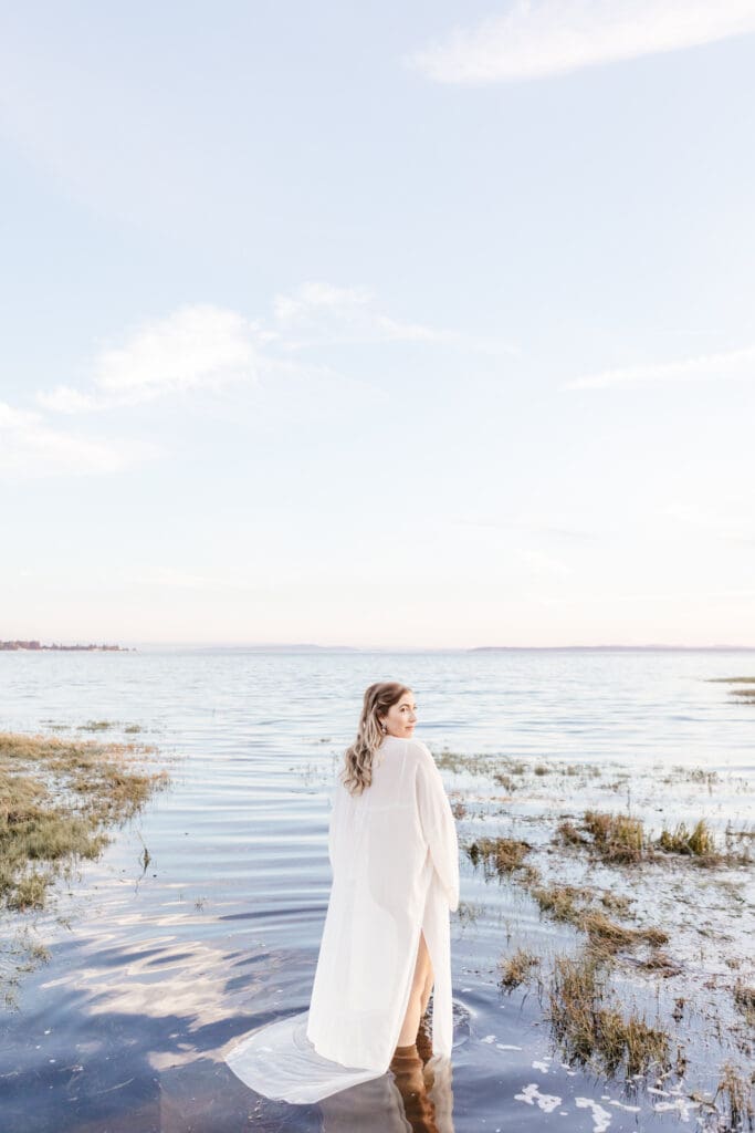 Woman in a simple white maternity dress standing barefoot near calm water