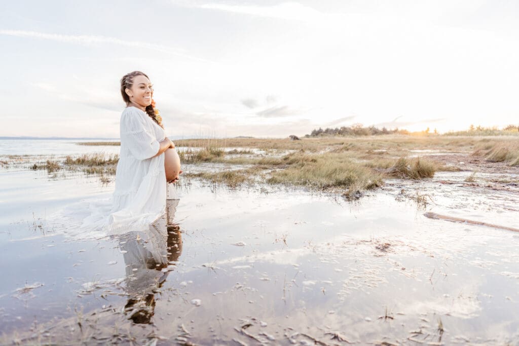 Pregnant woman standing in shallow water wearing a light, flowing maternity gown