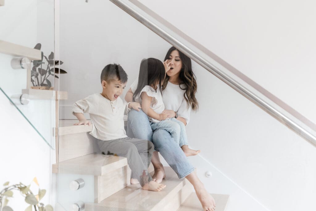 Children sitting on the stairs while their mother makes a silly face to make them laugh.
