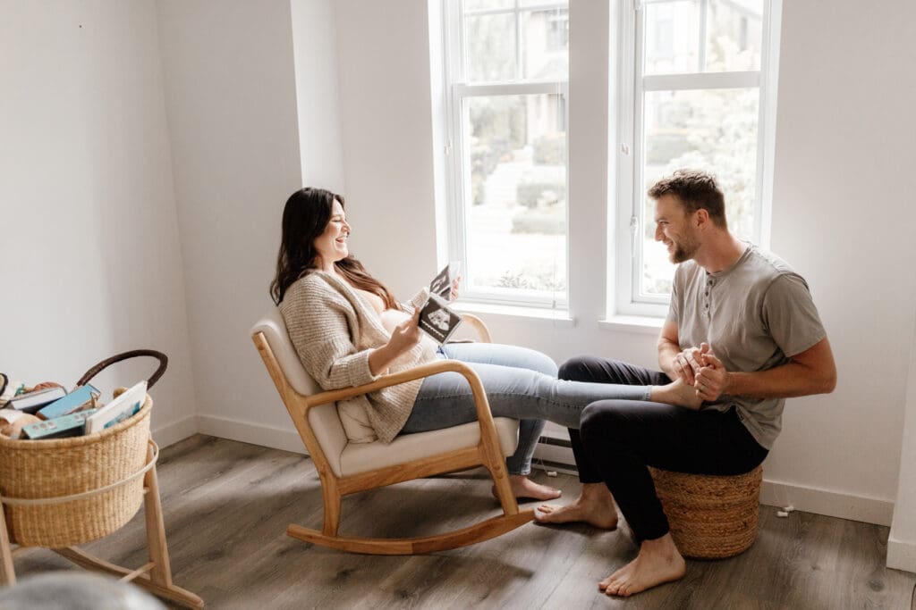 Pregnant woman and husband sitting by a window looking at their sonogram while she gets a foot rub during a calm in-home maternity photo session