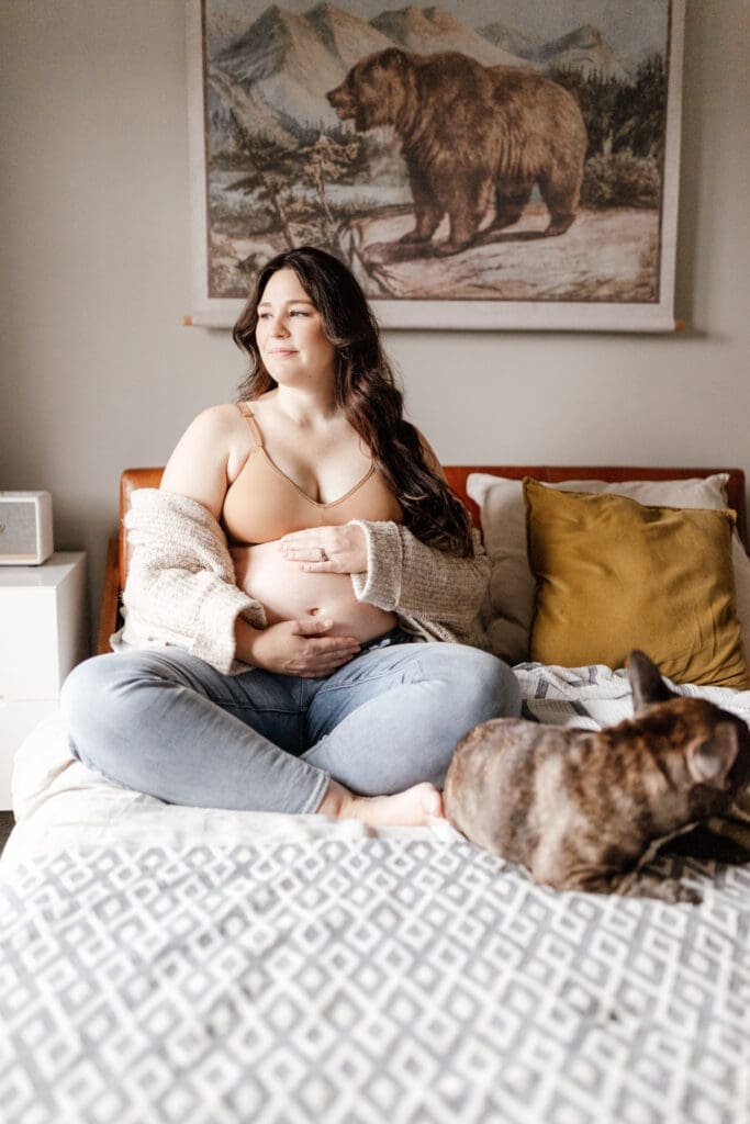 Expectant mother relaxing on a bed wearing a neutral sweater and jeans for an in-home maternity session