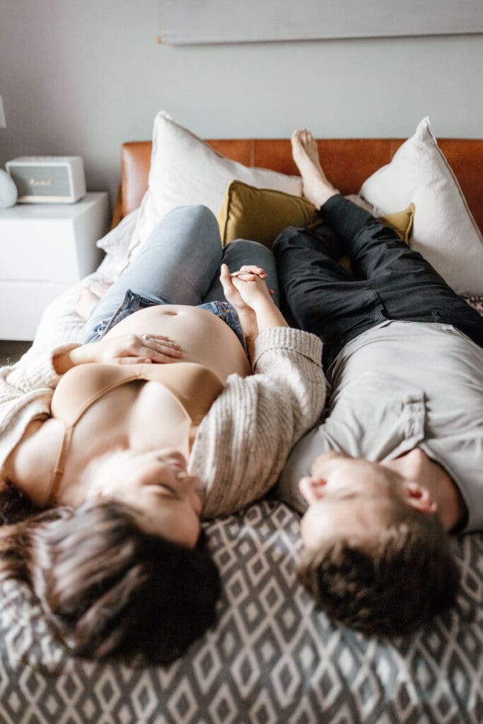 Couple lying on a bed during an in-home maternity session wearing comfortable neutral clothing