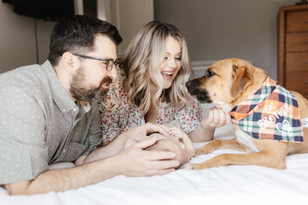 Parents lying on a white bed gently touching their newborn during an in-home session