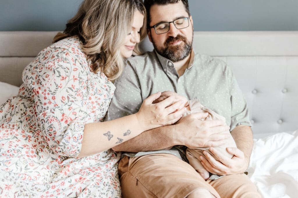 Parents lying on a white bed gently touching their newborn during an in-home session