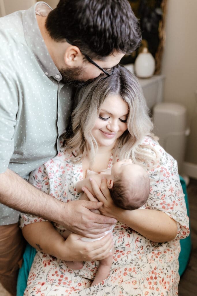 Parent holding newborn upright near a window while wearing coordinating outfits during their newborn photo shoot