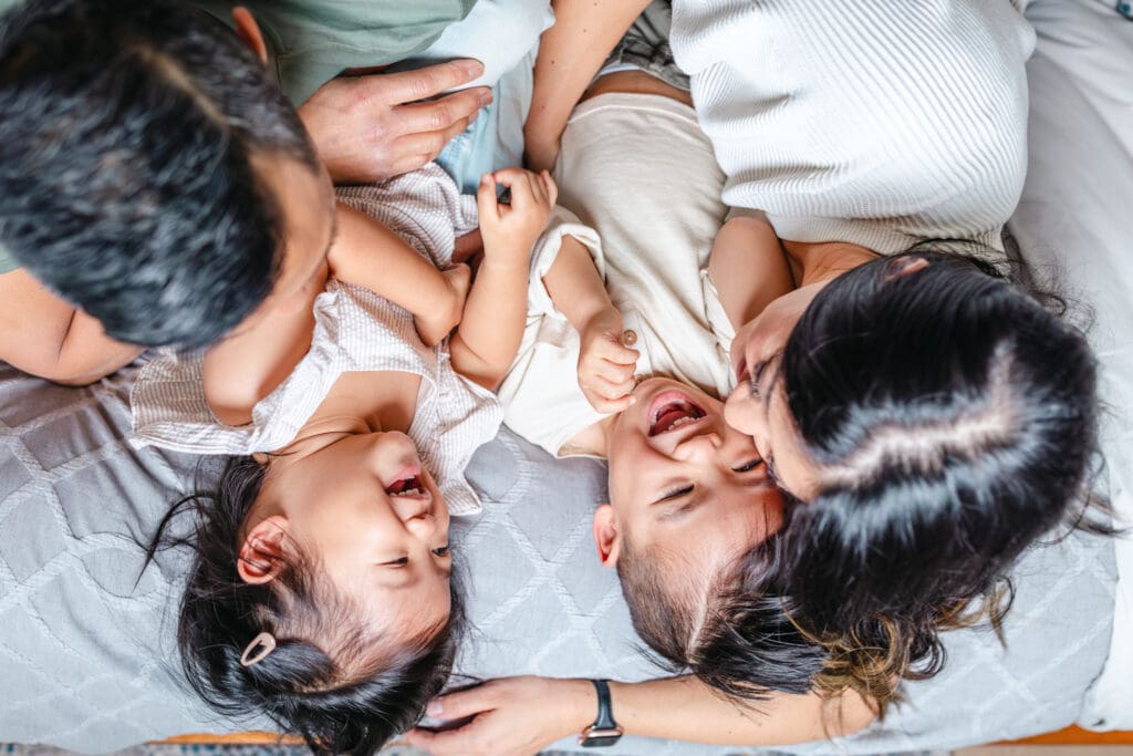Children giggling while lying close together on the bed with their parents’ arms around them.