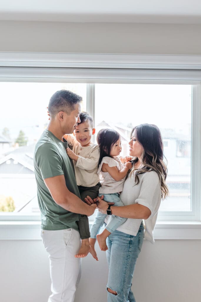 Family of four standing by a bright window, parents holding their children.