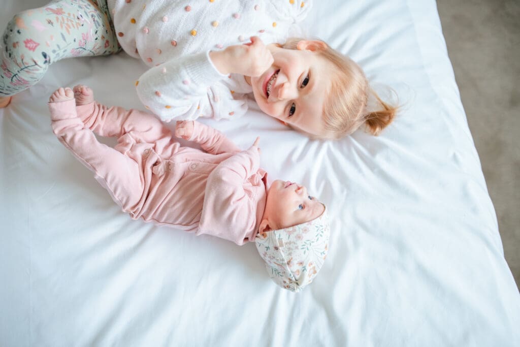 In-home family photo showing toddler smiling on white bed with newborn sister in a light and airy newborn photos