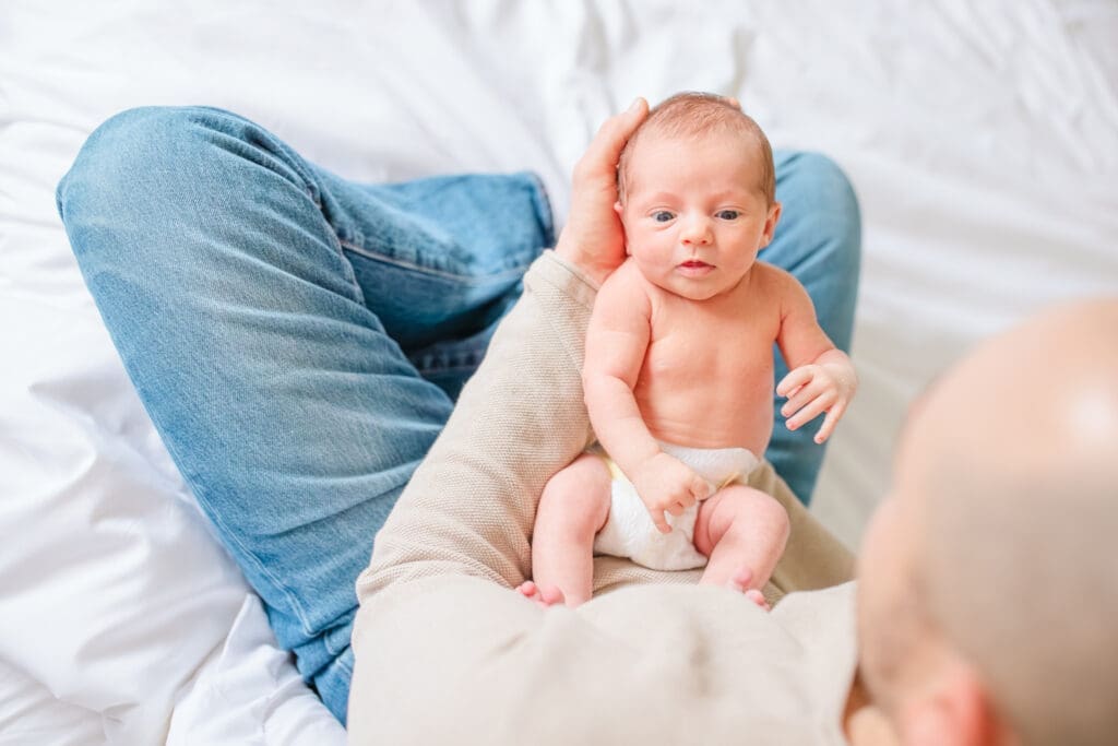 Father and newborn child. Dad is wearing soft neutral outfit with jeans and newborn in a diaper during an in-home family photography session
