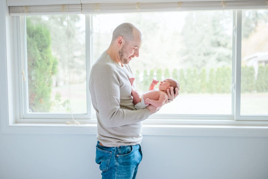 Father and newborn child standing by large picture window. Dad is wearing soft neutral outfit with jeans and newborn in a diaper during an in-home family photography session
