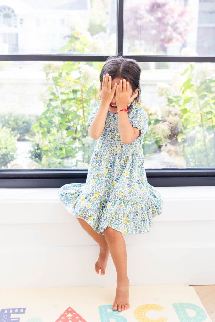 Young child sitting by a window during an in-home family photography session in Vancouver while playing peek-a-boo.